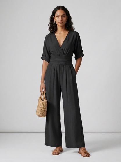 A woman modeling a black wrap-style jumpsuit with short sleeves, a cinched waist, and wide legs, holding a small woven straw handbag against a light gray studio background.