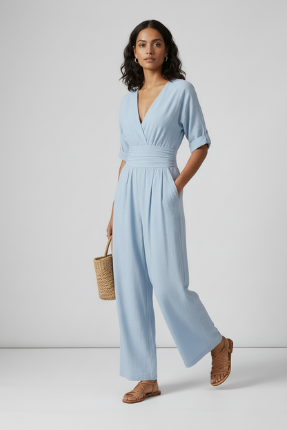 A woman stands against a light gray studio background wearing a light blue wide-leg jumpsuit with short sleeves, a wrap-style V-neck, and side pockets. She is holding a small woven tan bag and wearing tan sandals.