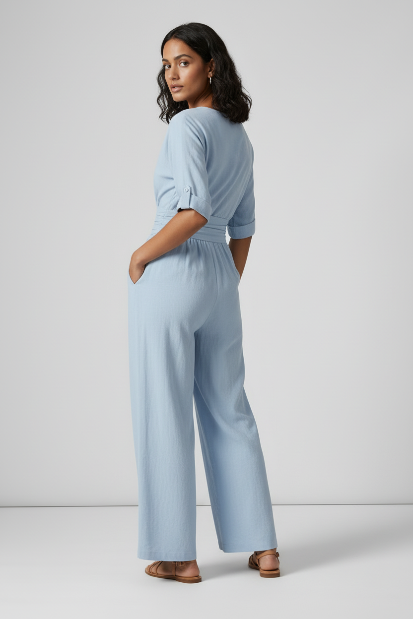 A woman stands against a light gray studio background wearing a light blue wide-leg jumpsuit with short sleeves, a wrap-style V-neck, and side pockets. She is holding a small woven tan bag and wearing tan sandals.