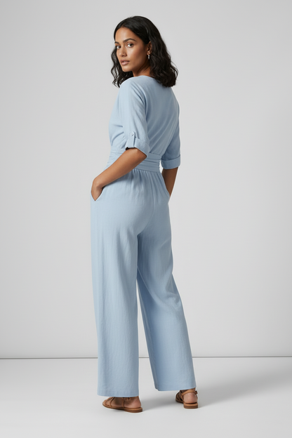 A woman stands against a light gray studio background wearing a light blue wide-leg jumpsuit with short sleeves, a wrap-style V-neck, and side pockets. She is holding a small woven tan bag and wearing tan sandals.