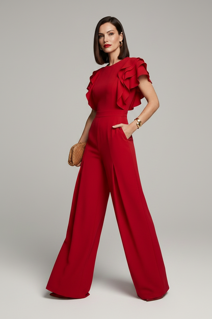 A woman modeling a bright red wide-leg jumpsuit with ruffled short sleeves, standing against a neutral light gray studio background.