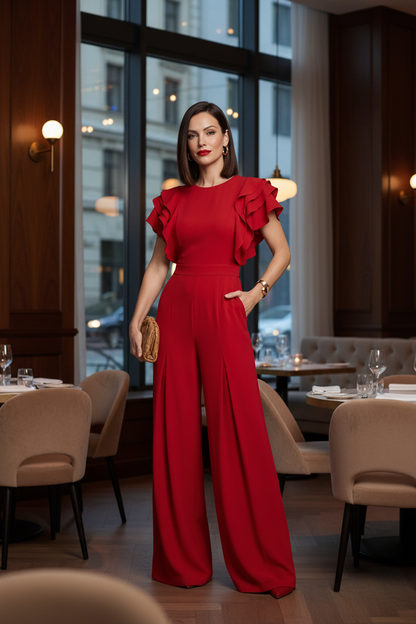 A woman modeling a bright red wide-leg jumpsuit with ruffled short sleeves, standing against a neutral light gray studio background.