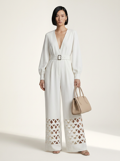 A model stands against a warm off-white studio background wearing a white long-sleeve wide-leg jumpsuit with a V-neck, matching belt, and geometric cutout detailing on the lower legs. She is holding a beige handbag.
