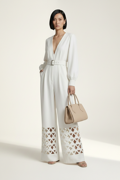 A model stands against a warm off-white studio background wearing a white long-sleeve wide-leg jumpsuit with a V-neck, matching belt, and geometric cutout detailing on the lower legs. She is holding a beige handbag.