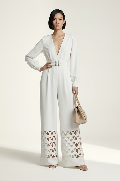 A model stands against a warm off-white studio background wearing a white long-sleeve wide-leg jumpsuit with a V-neck, a matching belt, and geometric cutout detailing on the lower legs. She is holding a beige handbag.
