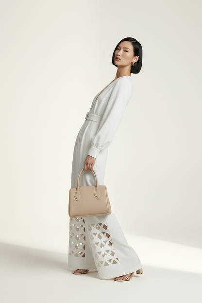 A model stands against a warm off-white studio background wearing a white long-sleeve wide-leg jumpsuit with a V-neck, matching belt, and geometric cutout detailing on the lower legs. She is holding a beige handbag.
