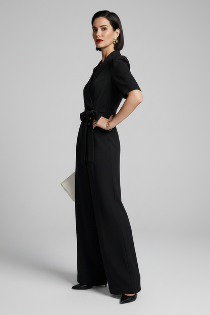A woman modeling a black short-sleeve wrap-style jumpsuit with a tied waist and wide legs, standing against a light gray studio background and holding a small white clutch.