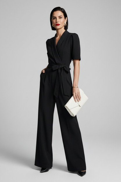 A woman modeling a black short-sleeve wrap-style jumpsuit with a tied waist and wide legs, standing against a light gray studio background and holding a small white clutch.