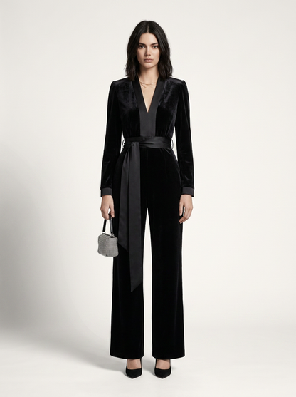 A model stands against a light gray studio background wearing a black velvet long-sleeve jumpsuit with a matching waist tie and wide legs, paired with black heels and a small gray handbag.