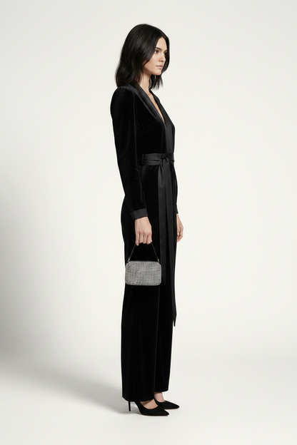 A model stands against a light gray studio background wearing a black velvet long-sleeve jumpsuit with a matching waist tie and wide legs, paired with black heels and a small gray handbag.