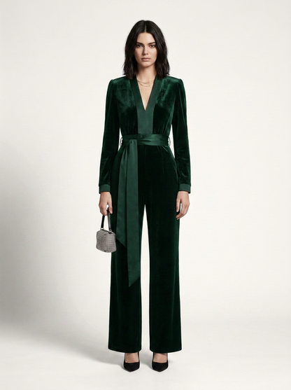 A model stands against a light gray studio background wearing a dark green velvet long-sleeve jumpsuit with a matching waist tie and wide legs, paired with black heels and a small gray handbag.