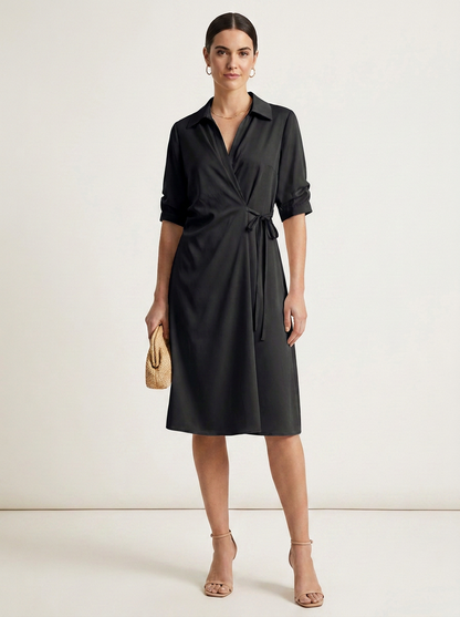 A woman modeling a black wrap-style midi dress with a collar, three-quarter sleeves, and a self-tie waist, standing against a plain light studio background. She is holding a small beige woven clutch and wearing nude high-heel sandals.