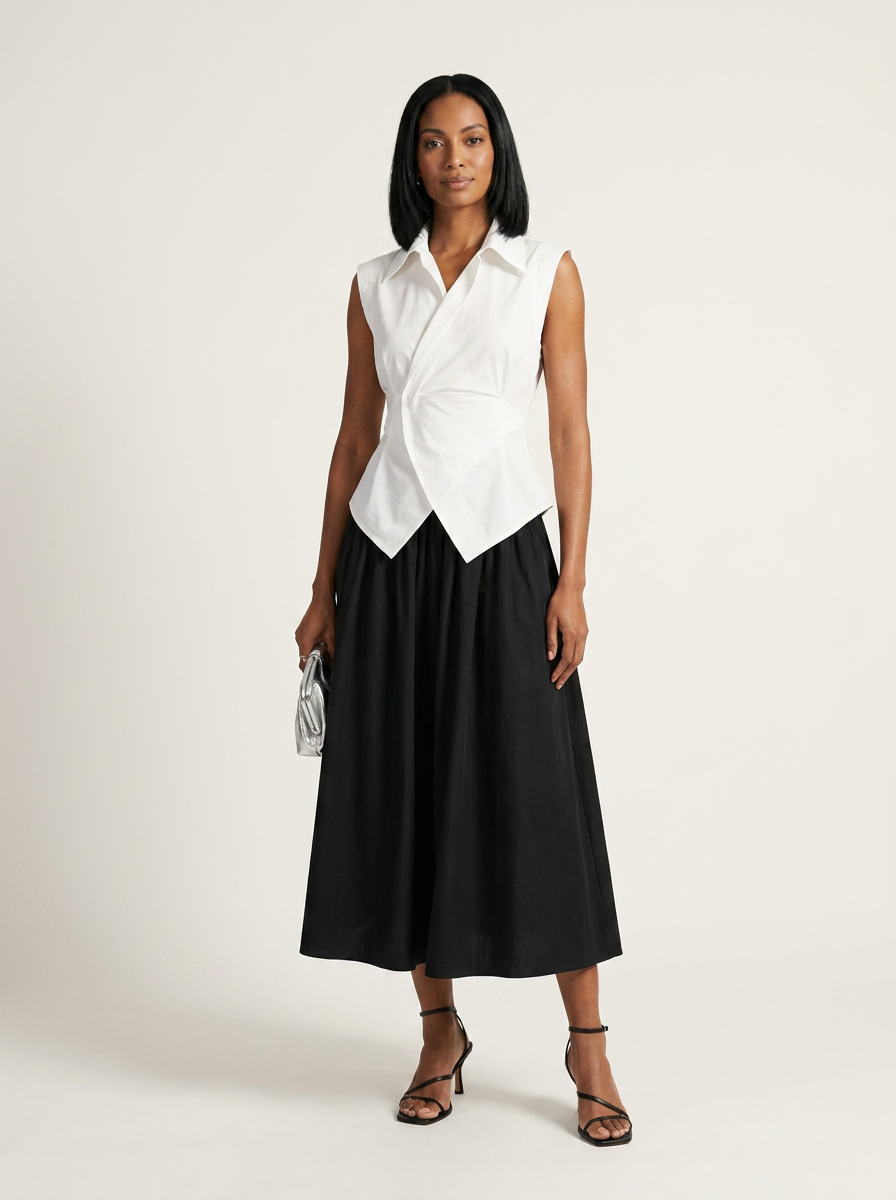 A model stands against a plain light beige studio background wearing a sleeveless white wrap-style shirt and a black midi skirt, holding a small metallic clutch.