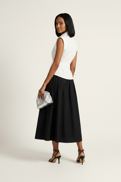 A model stands against a plain light beige studio background wearing a sleeveless white wrap-style shirt and a black midi skirt, holding a small metallic clutch.