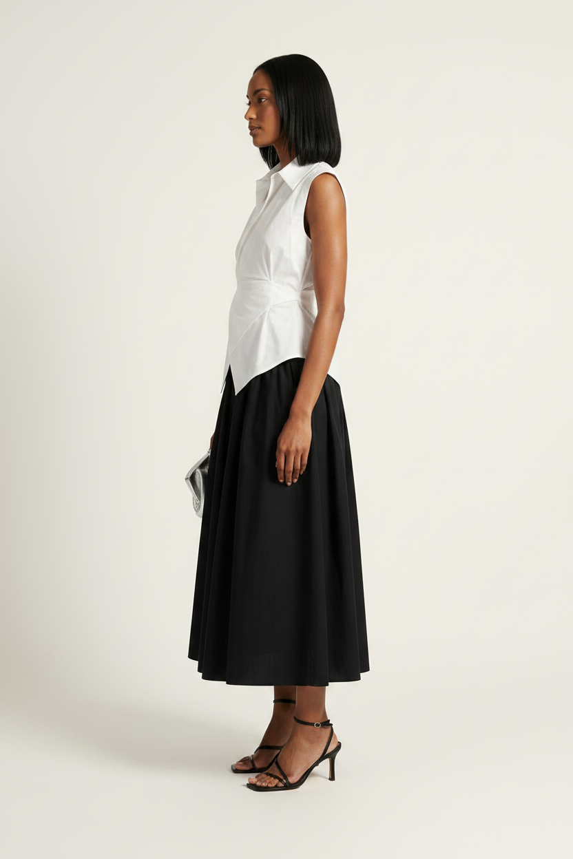 A model stands against a plain light beige studio background wearing a sleeveless white wrap-style shirt and a black midi skirt, holding a small metallic clutch.