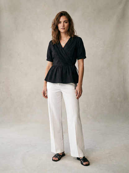 A woman stands against a textured light gray studio backdrop wearing a black short-sleeve wrap-style peplum top, white wide-leg pants, and black sandals.