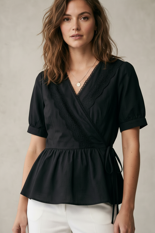 A woman wearing a black short-sleeve wrap-style blouse with lace trim and a peplum hem, paired with white pants, against a neutral textured studio background.