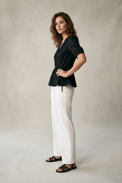 A woman modeling a black short-sleeve peplum wrap top with white wide-leg trousers and black strappy sandals, standing against a textured light gray studio backdrop.