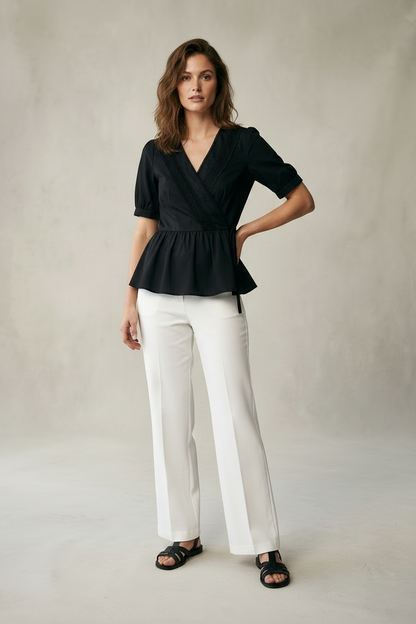 A woman modeling a black short-sleeve wrap-style peplum blouse with white wide-leg trousers and black sandals, standing against a neutral textured studio backdrop.