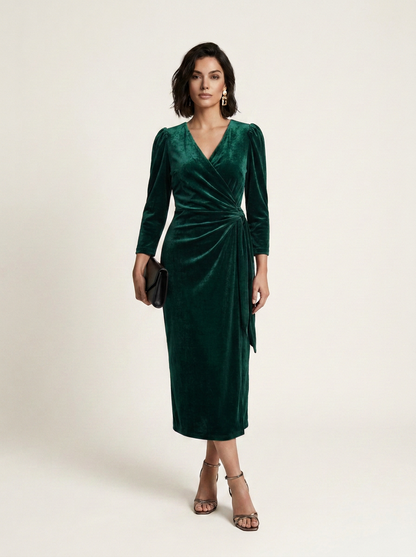 A woman models a dark green velvet midi wrap dress with long sleeves and a gathered waist, paired with brown strappy heels and holding a small black clutch against a plain light beige studio background.