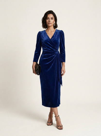A woman modeling a royal blue velvet midi wrap dress with long sleeves, a V-neckline, and a draped waist tie, paired with brown strappy heels and holding a small black clutch against a light beige studio background.