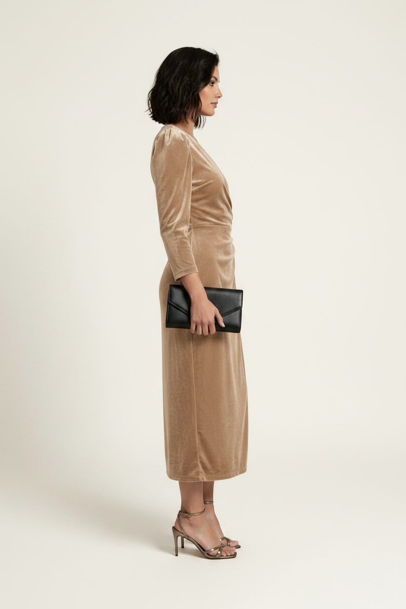 A woman models a long-sleeved beige velvet midi wrap dress with a draped waist and side tie, paired with brown strappy heels and holding a small black clutch, set against a plain light beige studio background.