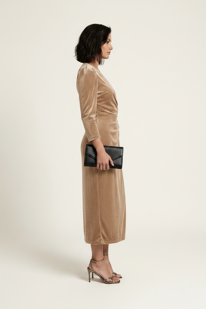A woman models a long-sleeved beige velvet midi wrap dress with a draped waist and side tie, paired with brown strappy heels and holding a small black clutch, set against a plain light beige studio background.