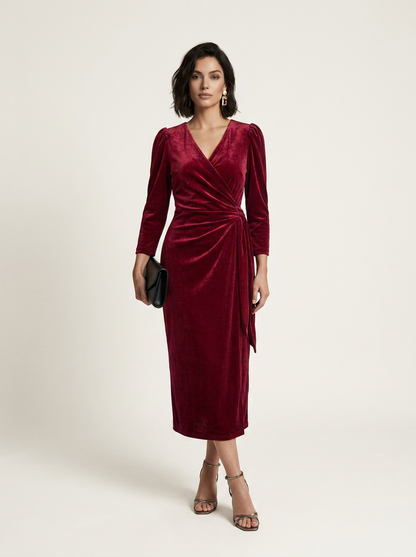 A woman modeling a long-sleeved burgundy velvet midi dress with a wrap-style V-neck and side tie detail, paired with metallic heeled sandals and a small black clutch, against a plain light beige studio background.